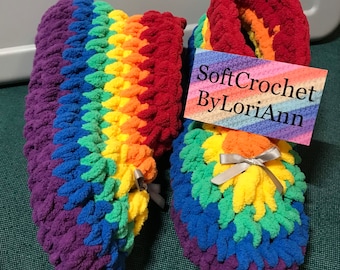 May include: A pair of rainbow-colored crocheted slippers with a purple base. The slippers have a fluffy texture and are tied with a gray ribbon. The text "SoftCrochet ByLoriAnn" is on a white tag.