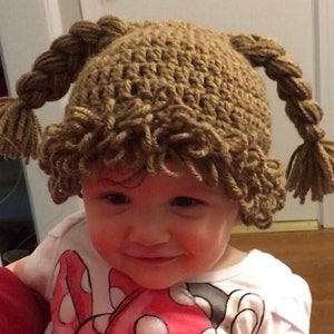 May include: A brown crocheted hat with two braided pigtails and a curly brown yarn wig. The hat is worn by a child with a white shirt and a red and white polka dot shirt underneath.