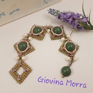 Peut inclure: Un bracelet en perles vertes et dor&eacute;es avec un motif carr&eacute; et des accents de pierres pr&eacute;cieuses vertes. Le bracelet est sur une surface blanche avec une fleur de lavande.