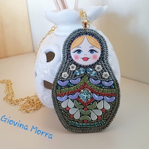 May include: A beaded necklace with a colourful Russian nesting doll pendant. The doll has a blue and white patterned dress, a green and red skirt, and a light blue and white floral design around the neck. The doll's face is painted with a pink blush, blue eyes, and a red mouth.