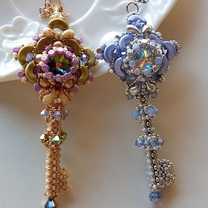 Könnte beinhalten: Zwei perlenbesetzte Anhänger in Schlüsselform, einer mit einem hellblauen und silbernen Design und der andere mit einem goldenen, violetten und grünen Design.