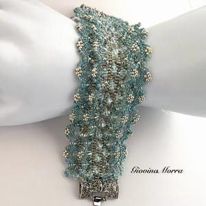 Può includere: Un braccialetto di perline nei toni del verde acqua e argento, con intricati motivi floreali. Il braccialetto ha una chiusura argentata ed è mostrato su una superficie bianca. Il nome del designer, Giovina Morra, è visibile. Il braccialetto misura circa 18 cm.