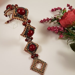 Peut inclure: Un bracelet en perles rouges et dor&eacute;es avec quatre grosses perles de pierres pr&eacute;cieuses rouges. Le bracelet est sur un fond blanc.