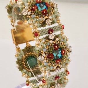 Könnte beinhalten: Ein Armband aus Perlen in einem grün-rot-goldenen Farbschema. Das Armband hat einen großen, quadratischen, türkisfarbenen Edelstein in der Mitte. Das Armband ist aus kleinen, runden Perlen gefertigt und hat einen goldenen Verschluss.