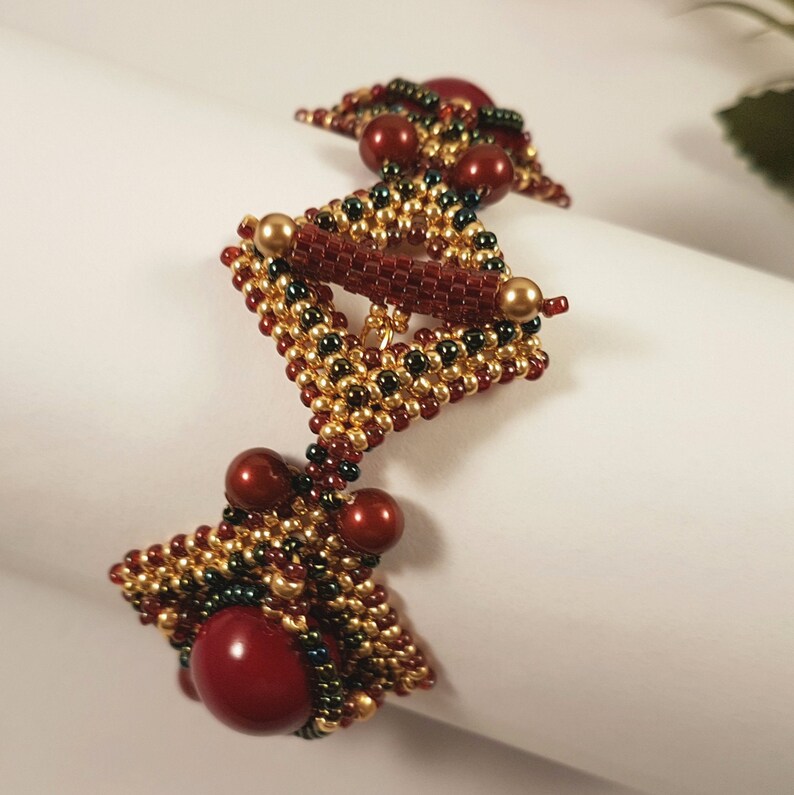 Peut inclure: Un bracelet en perles avec un motif g&eacute;om&eacute;trique dans les tons or, vert et rouge. Le bracelet pr&eacute;sente une forme triangulaire centrale avec des triangles plus petits de chaque c&ocirc;t&eacute;.