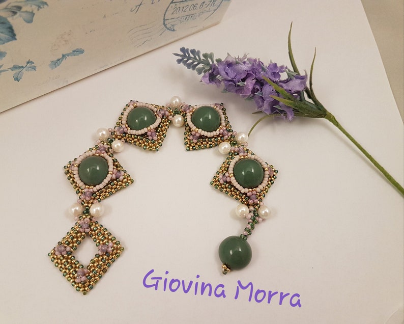 Peut inclure: Un bracelet en perles vertes et dor&eacute;es avec un motif carr&eacute; et des accents de pierres pr&eacute;cieuses vertes. Le bracelet est sur une surface blanche avec une fleur de lavande.