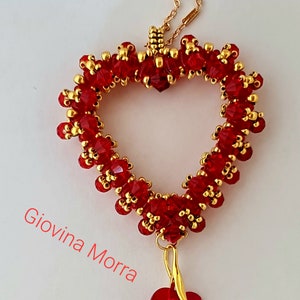 Può includere: Una collana con catena dorata con un ciondolo a forma di cuore rosso perlato. Il cuore è fatto di piccole perle rosse e dorate. Un cuore rosso più piccolo pende dalla parte inferiore del ciondolo.