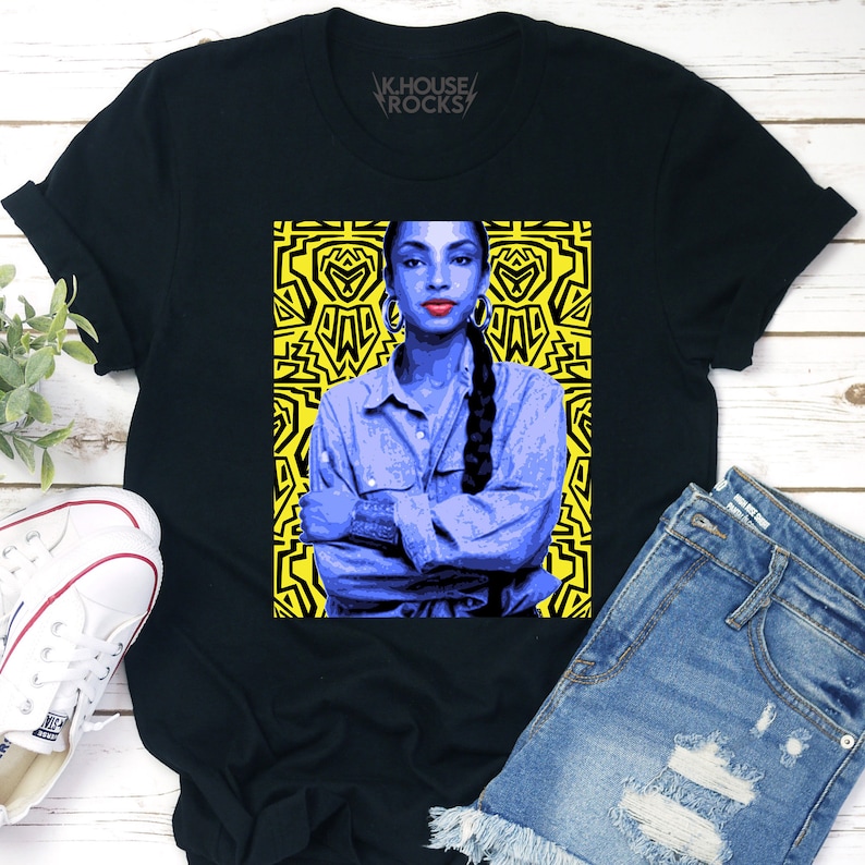 May include: Black t-shirt with a graphic of a woman with a blue shirt and long braid, against a yellow background with a geometric pattern. The text "KHOUSE ROCKS" is printed on the shirt.