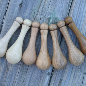 Puede incluir: Colección de siete herramientas de masaje de madera, cada una con un extremo bulboso y una parte superior redondeada. Las herramientas varían en color, desde tonos de madera claros a oscuros. Están dispuestas sobre una superficie de madera gris envejecida.