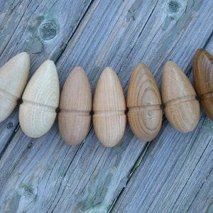 Puede incluir: Siete objetos de madera con forma de bellota en tonos marrones y beige, dispuestos sobre una superficie de madera gris envejecida. Cada bellota presenta un patrón de vetas distintivo y una banda tallada en el centro.