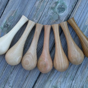 May include: Seven wooden spoons with teardrop-shaped bowls and long, slender handles. The spoons vary in colour from light beige to dark brown, showcasing the natural wood grain. They are arranged on a weathered, grey wooden surface.