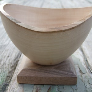 May include: A handcrafted wooden bowl with a light-colored interior and a darker rim, set on a square, dark-stained wooden base. The bowl's shape is gently curved, showcasing the natural wood grain. The base is approximately 4 inches square.