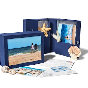 May include: A blue box with a photo of a beach scene and two people walking on the sand. The box is open and there are seashells and postcards inside.