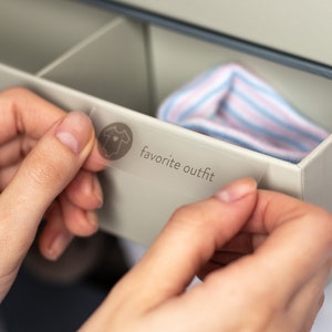 May include: A hand is placing a clear label with a baby onesie icon and the text "favorite outfit" on a drawer in a white dresser.