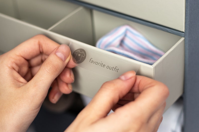 May include: A hand is placing a clear label with a baby onesie icon and the text "favorite outfit" on a drawer in a white dresser.