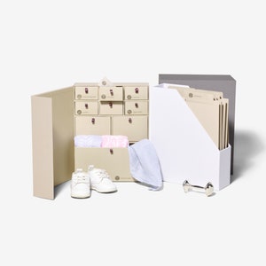 May include: A beige gift box with a drawer system containing various items, including a pair of white baby shoes, a blue towel, and a silver rattle. The box is open and the drawers are labeled with the words "Hospital Preparation", "Baby Essentials", "First Outfit", "Diaper Bag", "Going Home", and "Digital Prints".