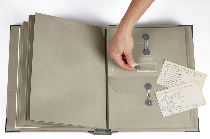 May include: A gray recipe binder with a clear plastic pocket labeled "savor" and a handwritten recipe card inside. The binder has a number "7" on the front and is open to reveal a clear plastic pocket on the right side.