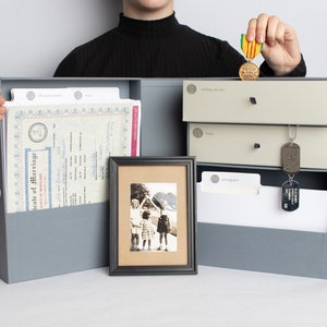 May include: A gray storage box with labeled compartments and drawers, holding documents, a framed photo, and military dog tags. A medal is held above the box. The box is designed for organizing keepsakes and important papers.
