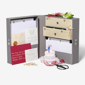 May include: A gray wedding keepsake box with three drawers for storing wedding mementos. The box is open and the drawers are pulled out, revealing compartments labeled "engagement", "wedding", and "something old, something new". The box also includes a small cake topper with a bride and groom, a red ribbon, and a pair of scissors.