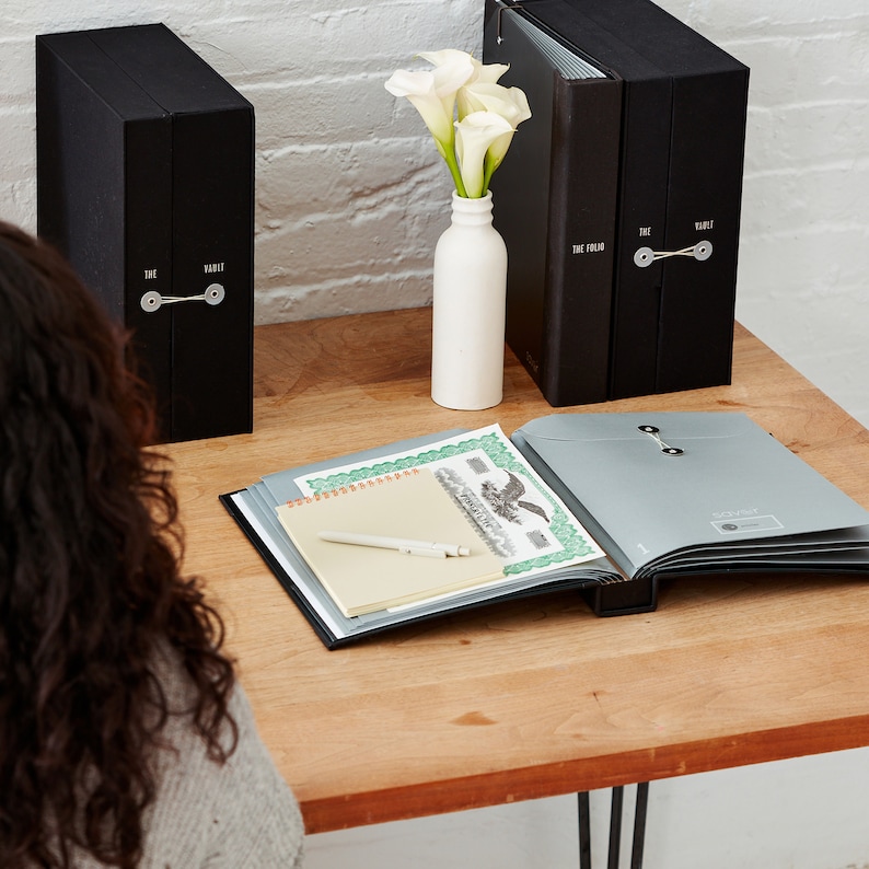 May include: A wooden desk with two black binders labeled "The Folio" and "The Vault", a white vase with white calla lilies, a silver folder with a black spine, a yellow notepad, and a white pen.