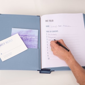 May include: A blue binder with a white page open to a table of contents. The page is labeled "The Folio" and has a handwritten name "Lucy's Bat Mitzvah". The table of contents lists "Invitations", "Programs", and "Venues".