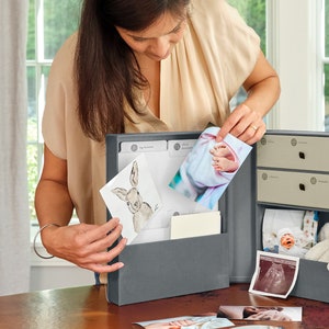 May include: A gray keepsake box with a hinged lid and two drawers. The box is open and filled with photos and mementos, including a photo of a baby's feet, a drawing of a bunny, and a sonogram. The box is a great way to store and organize precious memories.