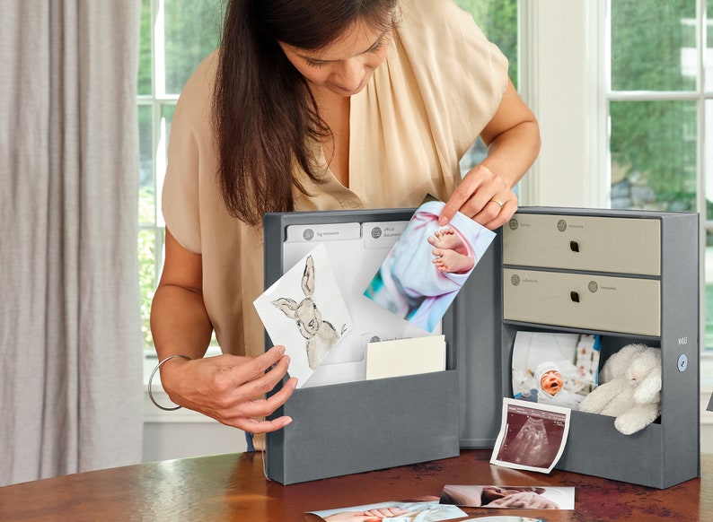 May include: A gray keepsake box with a hinged lid and two drawers. The box is open and filled with photos and mementos, including a photo of a baby's feet, a drawing of a bunny, and a sonogram. The box is a great way to store and organize precious memories.