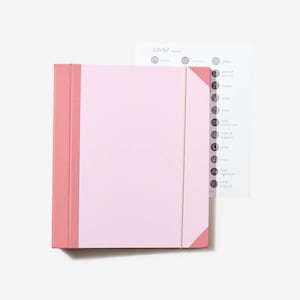 May include: A pink binder with a white label and a white elastic band. The binder has a pink spine and a pink flap on the right side. The label has the word "savoir" printed on it. The binder is sitting on a white surface.