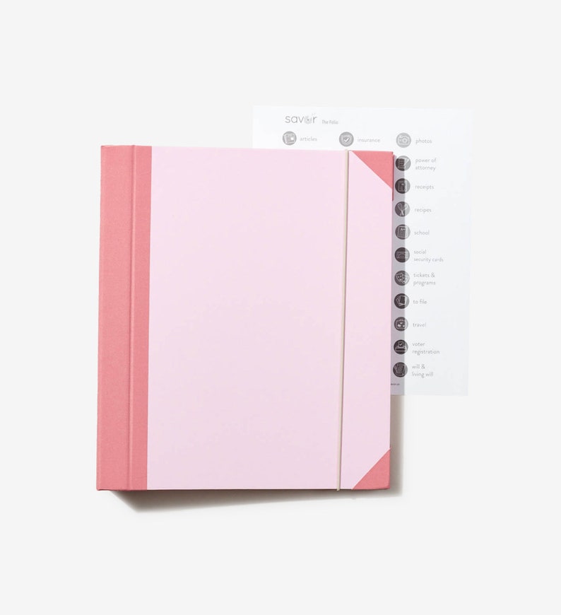 May include: A pink binder with a white label and a white elastic band. The binder has a pink spine and a pink flap on the right side. The label has the word "savoir" printed on it. The binder is sitting on a white surface.