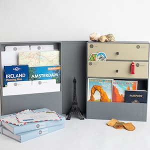 May include: A gray travel organizer with a drawer system for storing travel documents, maps, and souvenirs. The organizer has a built-in compartment for storing passports, tickets, and other travel essentials. The organizer is open and shows a variety of travel items, including maps, postcards, and a passport.