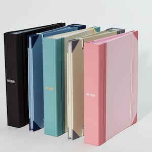 May include: Five different colored binders with the text "The Fold" printed on the front. The binders are arranged in a row from left to right, with the black binder on the left and the pink binder on the right.