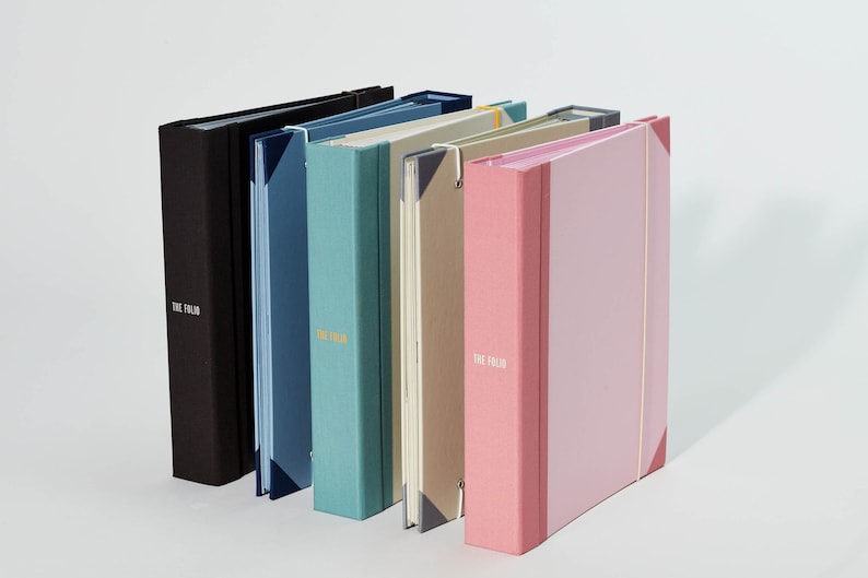 May include: Five different colored binders with the text "The Fold" printed on the front. The binders are arranged in a row from left to right, with the black binder on the left and the pink binder on the right.