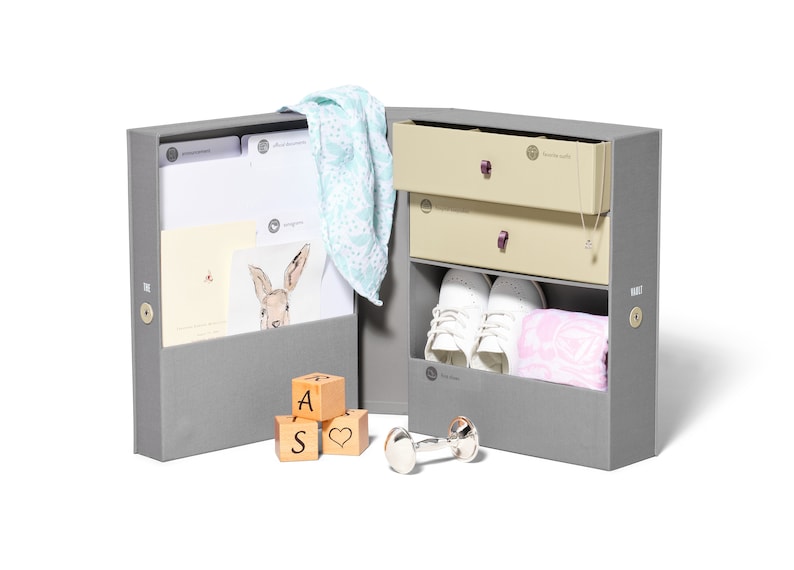 May include: A gray keepsake box with a light blue blanket, wooden blocks that spell "A S" and a heart, a silver rattle, and a pair of white baby shoes. The box has two drawers with a beige interior and a purple pull.