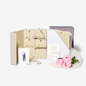 May include: A set of wedding stationery supplies in a beige box with drawers. The box contains a card with a photo of a couple, a card with the text "Caroline Davis & Daniel James Baker", a white folder with beige dividers, and a bouquet of pink peonies.