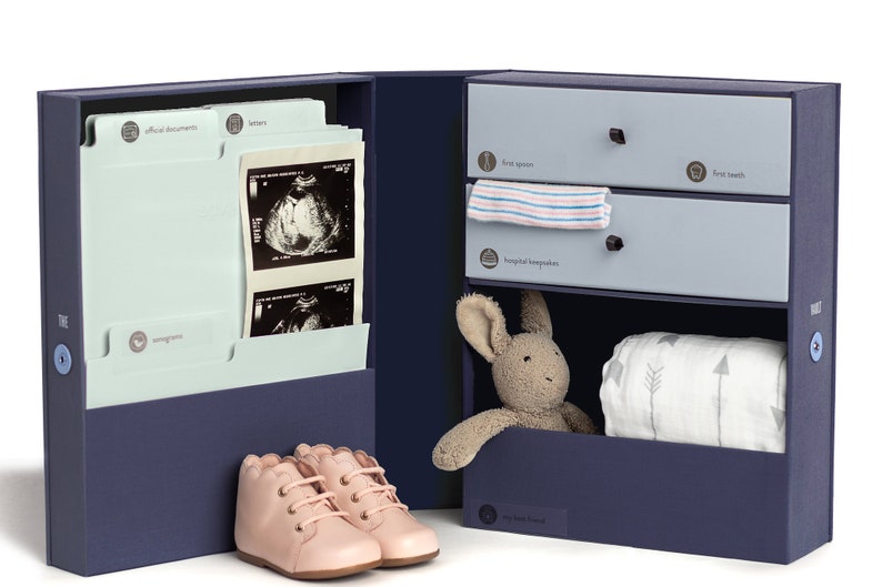 May include: A navy blue keepsake box with a light blue interior. The box has a drawer with a label that says "first spoon" and another drawer with a label that says "first teeth". The box also has a shelf with a label that says "hospital keepsakes". The box is open and there is a stuffed bunny rabbit, a baby blanket, and a pair of pink baby shoes inside.
