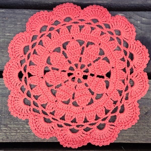 May include: A coral pink crocheted doily with a circular design, featuring intricate floral patterns. The doily is laid flat on a wooden surface.