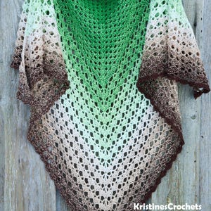 May include: A handmade crochet shawl in a V-shape, featuring a gradient of colors from green to brown. The shawl has an open, lacy design and is displayed against a wooden background. The shawl is a wearable accessory.