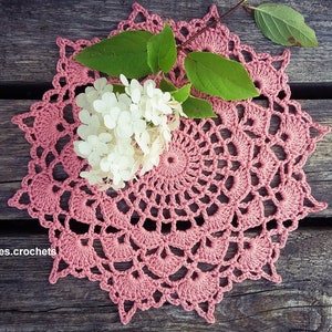 May include: A pink crocheted doily with a delicate floral pattern. The doily is laid on a wooden surface with a white flower on top.