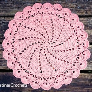 May include: A light pink, circular crochet doily with an intricate, openwork pattern and a scalloped edge. The text "KristinesCrochets" is visible. The doily is displayed on a dark wooden surface. The doily is a decorative item.