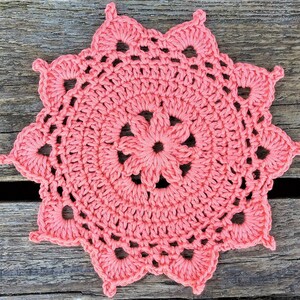 May include: A pink crocheted doily with a starburst design. The doily is made with a delicate, lacy stitch and has a circular shape.