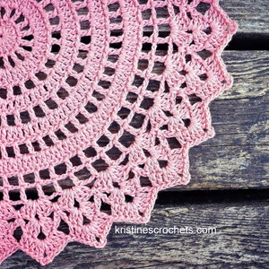 May include: A pink crocheted doily with a delicate, intricate design. The doily is laid on a weathered wooden surface.