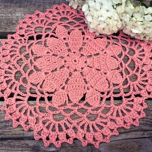 May include: A delicate pink crochet doily with a floral design, resting on a weathered wooden surface.