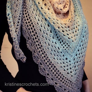 May include: A crocheted shawl with a gradient of colors from light blue to beige. The shawl is made with a delicate stitch pattern and has a fringed edge.