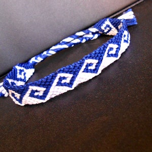May include: A blue and white friendship bracelet with a geometric pattern. The bracelet is made of woven threads, with a repeating design of blue squares and white spaces. The bracelet is lying on a dark surface.