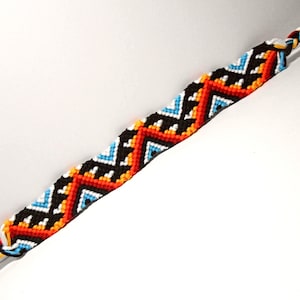 May include: A woven friendship bracelet with a geometric pattern in a chevron design. The bracelet features black, red, orange, white, and blue colours. The ends are braided with multiple colours.