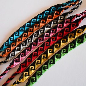 Può includere: Una collezione di braccialetti dell'amicizia fatti a mano in vari colori. Ogni braccialetto presenta un motivo geometrico ripetuto in nero, con accenti di turchese, arancione, bianco, rosa, rosso, giallo e verde. I braccialetti sono esposti su una superficie bianca.