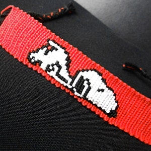 Peut inclure: Bracelet d'amitié rouge et noir fait à la main avec un motif Snoopy blanc et noir. Le bracelet est fait de fils tissés et possède des liens tressés noirs pour la fermeture. Un accessoire unique.
