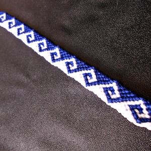 May include: A handmade friendship bracelet featuring a repeating pattern of blue and white geometric shapes. The bracelet is made of woven thread and is displayed against a dark background.