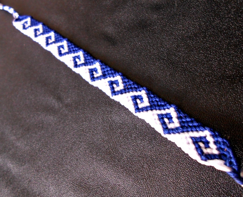 May include: A handmade friendship bracelet featuring a repeating pattern of blue and white geometric shapes. The bracelet is made of woven thread and is displayed against a dark background.