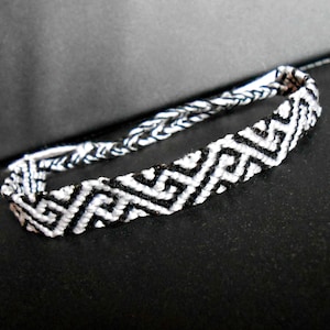 Könnte beinhalten: Schwarz-weiß gewebtes Freundschaftsarmband mit geometrischem Muster. Das Armband hat ein sich wiederholendes Zickzack-Design in Schwarz und Weiß, das einen kräftigen Kontrast erzeugt. Das Armband besteht aus gewebten Fäden.